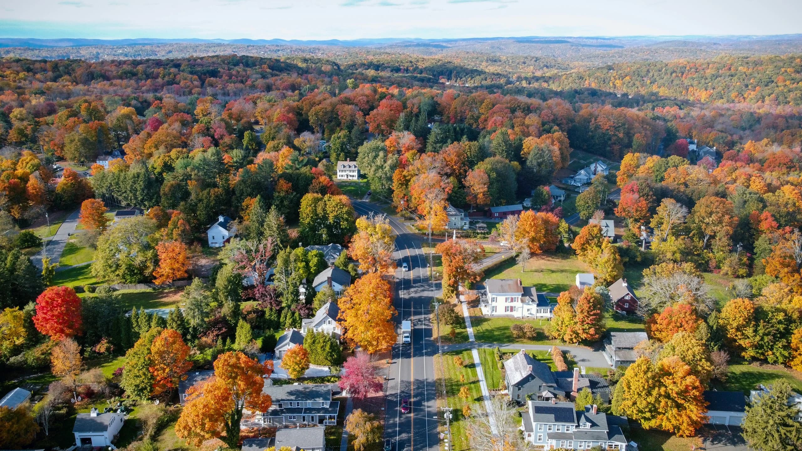 Aerial view over a Connecticut village in autumn — white clapboard houses and a red barn along a two-lane road, under hardwood canopy in full colour, with wooded hills to the horizon