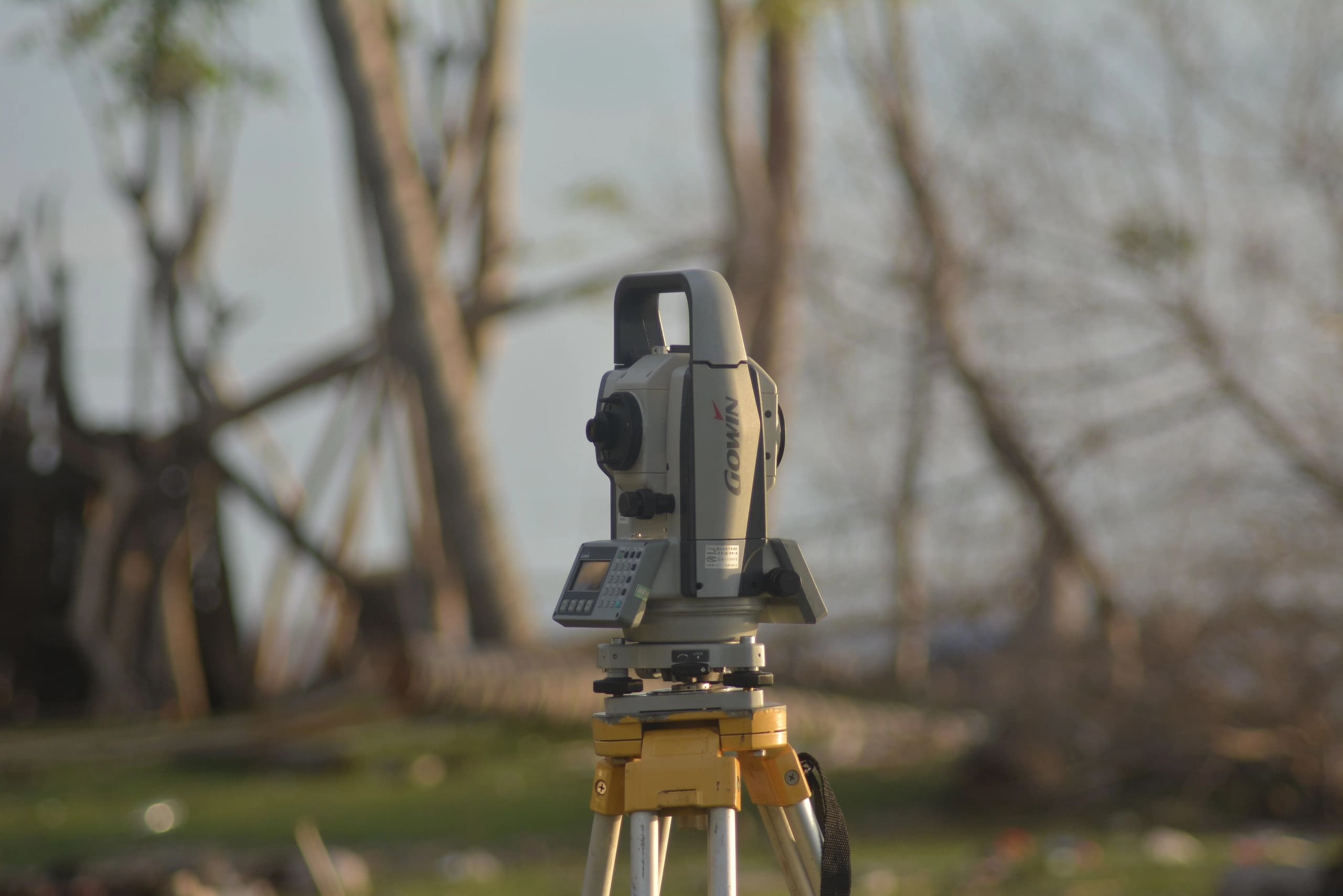 Total station mounted on a tripod at the edge of a wooded lot at dusk