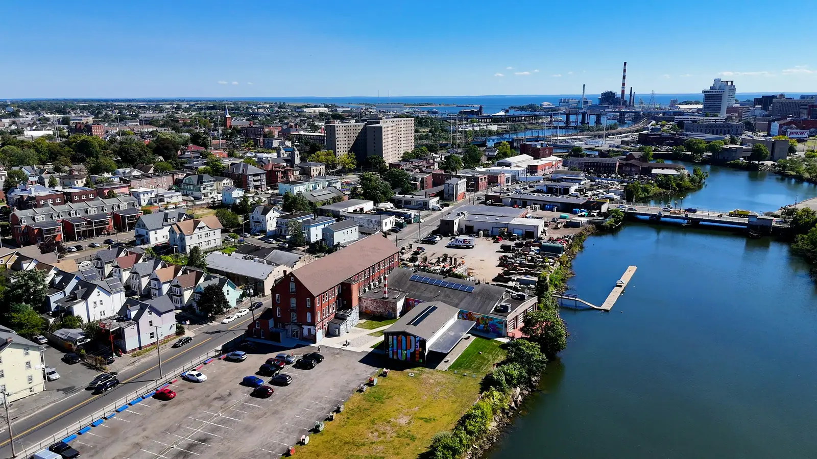 Aerial view of a waterfront commercial district, brick blocks and yards along a river with bridges and port cranes beyond